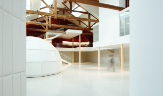 Photographie représentant un espace d’exposition illuminé par la lumière du jour, avec un sol blanc et des murs blancs. Un igloo géométrique blanc est au centre de l’espace. 